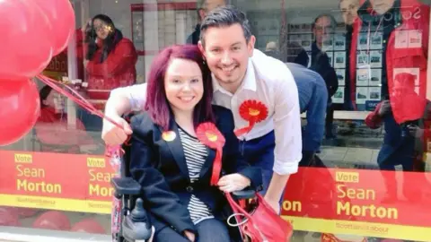 Facebook Pam Duncan-Glancy, who has shoulder length hair dyed red, in a wheelchair next to Sean Morton, who has short black hair. She is wearing a dark suit jacket and trousers, while he is wearing a white shirt. They are standing in front of a glass store front, with red Labour on it. Morton is holding red balloons, while Glancy holds a red handbag. 