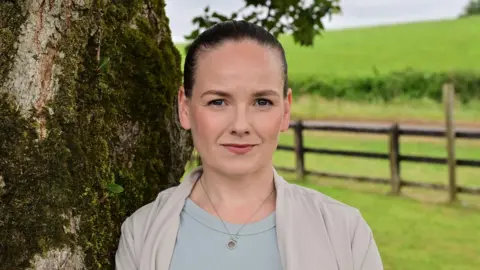 PA Media Anna Owens has black hair swept back from her forehead. She is standing beside a tree with fields in the background.