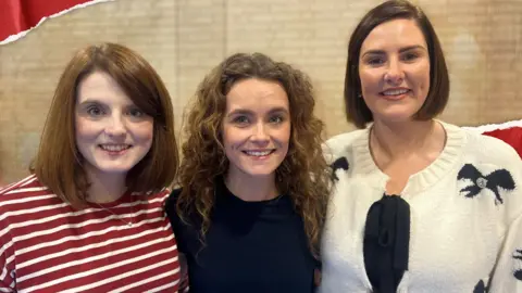 Toddler Sense North East's Jessica Brown (centre) a woman with long brown curly hair in a black top, pictured alongside Alex McNeill (left) and Fiona O'Connor (right) - both women have brown bob haircuts, Alex is in a striped burgundy and white top, Fiona is wearing a cream knitted cardigan with black bows - women are pictured against a cream brick wall 