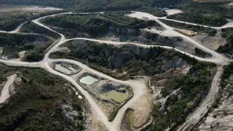 An aerial view shows a former china clay pit with pale, dusty tracks winding through excavated land and several small, still pools of greenish water. The site is surrounded by patches of scrub and trees, with exposed rock faces and industrial-looking clearings suggesting past or ongoing quarrying activity.
