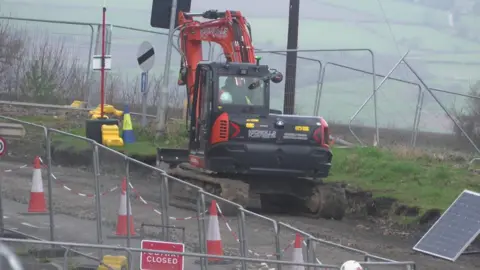 The image shows a roadworks scene on a rural road. An excavator is digging into the surface while surrounded by cones, fencing and temporary signs. Two workers in hard hats and high‑visibility clothing are standing nearby. A “closed” sign is placed in front of the work area. A solar‑powered panel is positioned to the right.
