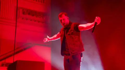 A lead singer on stage with arms outstretched taking the applause of fans. The stage lighting is read and he's wearing a denin waistcoat