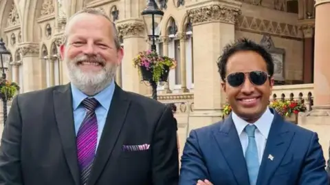 West Northamptonshire Council James Petter with short grey hair and beard, wearing a dark suit and purple tie and smiling at the camera. He is next to Zia Jusuf with short dark hair, wearing a blue suit and light blue tie and smiling at the camera. They are standing in front of Northampton's stone-built Guidhall