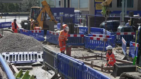 BBC Workers in high-visibility clothing carrying out water main repair work at a roadworks site, with excavators and blue safety barriers surrounding the excavation.
