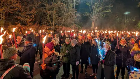 Participants of the torchlight parade walk through the streets of Lincoln. The majority of people are wearing warm, winter clothing and are carrying long candles which are burning. Trees are visible in the background.