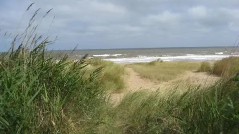 A view of the sea from the sand dunes on a cloudy day, but the sun is shining through. Tall patches of seagrass are blowing in the breeze and the waves are dark with white caps.