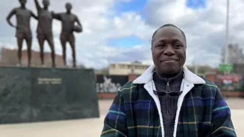 Lanre wearing a grey jumper and blue and green checked fleece with sunglasses smiling around his neck standing in front Manchester United's Holy Trinity statues.