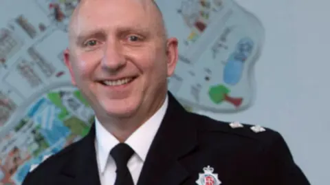 Jim Jones smiles as he is photographed in his police inspector's uniform. He wears the Greater Manchester Police shield.