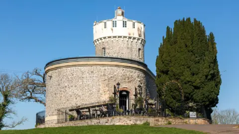 Getty Images The Clifton Observatory seen from outside. It is a circular stone building with a smaller circular tower at one end. There is an outdoor seating area with chairs and tables inside a dark metal fence, and a large conifer-style tree