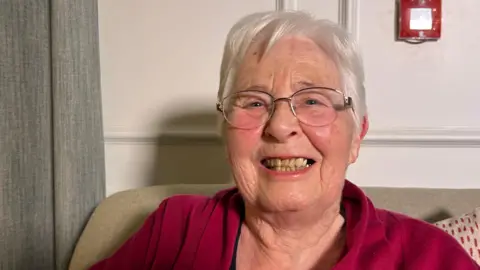 Joy, a 91-year-old woman with short white hair, smiles as she sits on a sofa in a care home. She is wearing glasses and a red fleece.