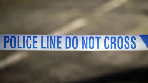 Blue and white police tape that reads 'police line do not cross'. A grey blurred road is behind it.