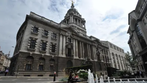 A view of the Old Bailey from the other side of the road near a water feature. The old part of the building can be seen with pillars and decorative windows and a statue of Lady Justice on the top.