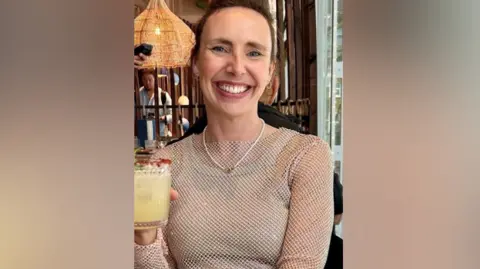 Meghan Lawson A smiling Rebecca Horne holding a glass with a pale yellow drink. She appears to be at a restaurant, a man an be seen at a table behind her. She is wearing a mesh top.