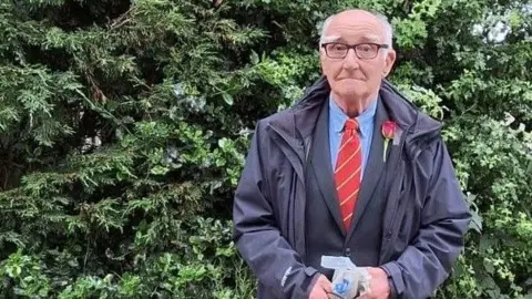Clare Wilson Christopher wears a red rose in his lapel and a dark suit with red tie. He wears a jacket over his suit and stands in front of greenery.