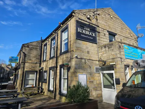 Aisha Iqbal/BBC The front of a traditional country pub. A black sign identifies The Roebuck pub. The building is Yorkshire stone, and has white windows with black trim. Banners hanging from the building indicate it is currently closed and new management is sought.
