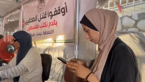 Two journalists working inside the Journalists Syndicate tent - one of them writes on a mobile phone, and the other sits in front of a large fan