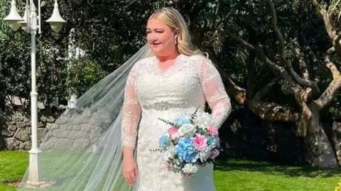 Kayley Stead A blonde-haired pistillate   wearing a achromatic  wedding formal  and holding a bouquet of white, pinkish  and bluish  flowers.