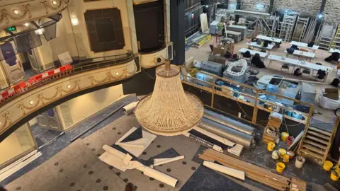 BBC Inside the Jersey Opera House during its recent £13m renovation. A chandelier is hanging from the ceiling. Lots of building materials are scattered on the floor and the venue's stage.