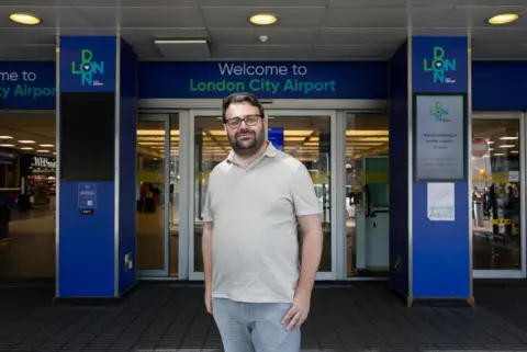 MyLondon A man in an off-white polo shirt and grey jeans standing in front of London City Airport entrance. He has dark brown hair, a beard and is wearing glasses. Behind him the entrance reads "Welcome to London City Airport". 