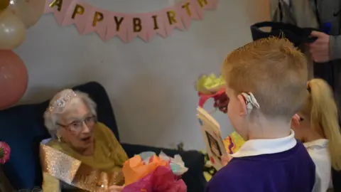 Percy Hedley Foundation Maisie Dalziel, who is wearing a sparkly sash and a tiara, looks pleasantly surprised as two children hand her gifts and cards.