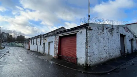 Lucy Blackledge Industrial unit on the corner of a small tarmac road. The unit is white and ha sa red metal shutter. There is barbed wire on the fence