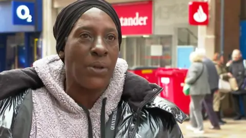 Monica is wearing a headscarf and fluffy jumper underneath a grey winter jacket. She is stood in a street in Wolverhampton.
