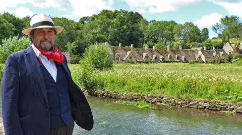 The Cotswold Explorer Mr Shuckburgh by a river on a sunny day. He is looking at the camera. There are old cottages on the other side of the bank, on a green meadow.