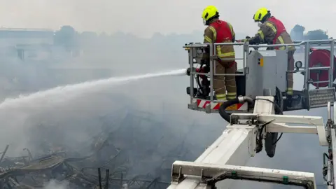 Two firefighters on an aerial platform working to tackle a blaze at a bathroom showroom. Smoke is coming off the building and is in the air. 