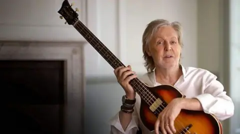 A recent photograph of Paul McCartney wearing a white shirt, seated in front of a fireplace and smiling into camera. He is holding a Hofner violin bass.