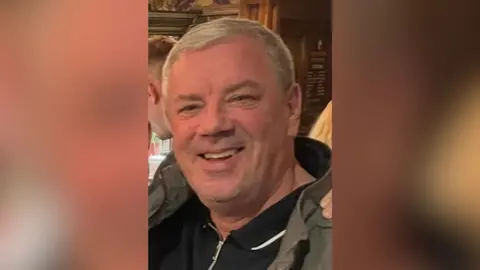 Gary Forbes A man with grey hair, wearing a green jacket and black polo shirt underneath is stood at what seems to be a bar or restaurant. He is smiling and looking directly at the camera