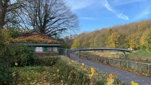 Durham County Council The old toilet block is a one story brown-brick building with a triangular roof, green door and window panes. There is a pile of yellow leaves on the roof. NO UNAUTHORISED ACCESS is written in red on a sign attached to the fenced-off building. There is a promenade next to it with the arched Baths Bridge visible in the distance. There are many trees across the river, with their leaves falling off. 