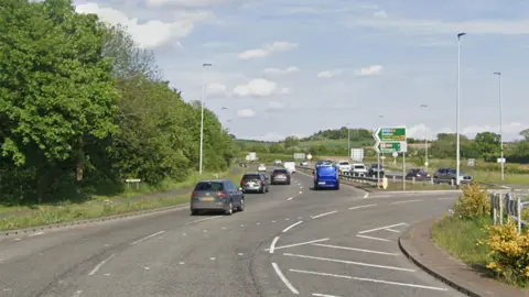 A roundabout on a dual carriageway with multiple vehicles exiting and a signpost pointing the way to Evesham and Stratford and a hospital
