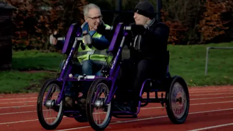 Two men, one on the left with grey hair and wearing glasses and another on the right wearing a hat, are sitting alongside each other on a type of bike, which is in part purple. It is on an athletics track and grass is in the background.