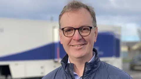 Surgeon Jonathan Ferguson wears black-rimmed glasses and has short grey hair. He is wearing a thin navy puffer jacket and a purple shirt. He is standing in front of a screening mobile unit.