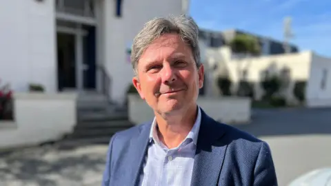 Deputy Andy Sloan - A man with grey and black hair smiling at the camera, wearing a blue jacket and a blue button up shirt. 
