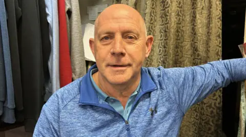 Christopher White in his shop. We can see clothes behind him. He is wearing a blue fleece and a blue polo underneath. He is clean shaven and has little hair.