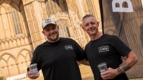 Sam Smith Sam Smith is wearing a baseball cap and a black T-shirt. He is standing next to Chris Coley who has short, fair hair and is also wearing a black top. Both are posing for the camera in front of Peterborough Cathedral.