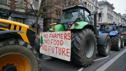 PA Media Four tractors in a line on a road in the middle of Westminster. A green tractor is in the middle of the image with a sign on the back that reads, "No farmers, No food, No future". A row of houses are visible in the background as well as a tree with no leaves.