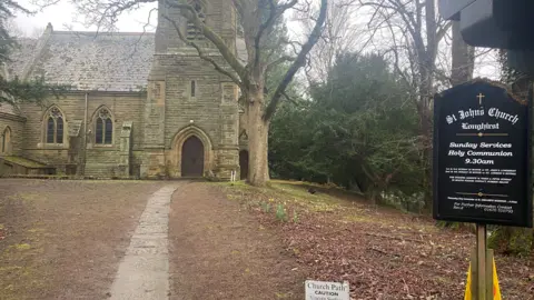 The outside of Longhirst Church, a medieval building with a steep spire. There is a board outside which gives times of the services.