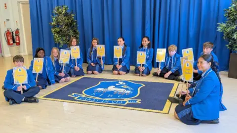 Handout A group of children in blue uniforms holding up yellow signs. On the floor is a blue rug with a yellow binding and school crest.