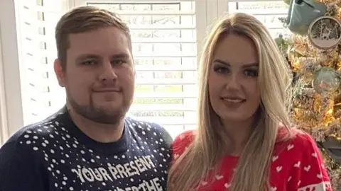 Ian wearing a navy sweater with white spots on and white writing which says "YOUR PRESENT IS UNDER THE TREE" in capital letters. He has short, light brown hair, a moustache, and a beard. He is standing with his arm around Jo Dayton, who is on the right. She has long, blonde hair and is wearing a red jumper with white love hearts and snowflakes on. Behind her on the right is a green Christmas tree decorated with sage green baubles, wooden decorations and white flowers. They are standing smiling at the camera in front of windows with white shutters, which are open. 