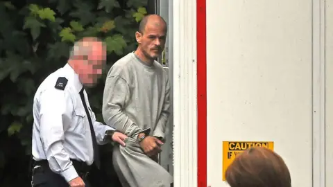 Pacemaker A man in grey prison sweats is handcuffed and is being guided into a white custody van by a prison officer wearing a white shirt, black tie and black trousers. The prison officer's face is blurred.