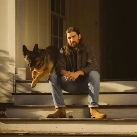 Patrick McCormack Noah Kahan poses on the porch of a rural house in America, as a dog bounds down the steps