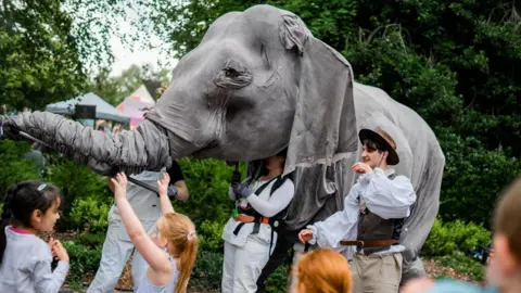 Jamie Bellinger A life-sized grey elephant puppet being controlled by a group of puppeteers through Bath Carnival