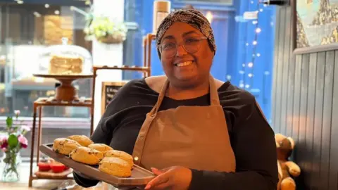 Woman in cafe holding scones up on a board. She has glasses and is wearing a blue top