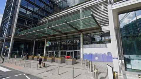 A general view of the outside of Brent Civic Centre. The Brent Council logo is in view alongside the writing of the name of the centre. There are people that can be seen in the image also.