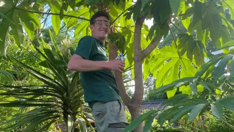 Dave Robinson Dave Robinson at the top of a tree in the couple's garden. He is stood to the side and smiling at the camera. In his hand is garden secateurs. He is surrounded by large green leaves. He is wearing protective eye goggles, a dark green polo top and khaki green shorts.
