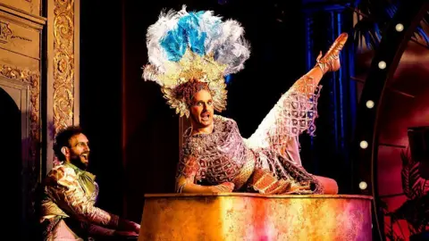 Pamela Raith Seiriol Davies is on top of a grand piano in a brown and gold dress, which gives the impression of a Roman soldier or Egyptian ruler. They wear an elaborate headdress of gold segments topped with peacock blue and white feathers. Another performer wearing a gold jacket sits playing the piano. Both appear to be singing.