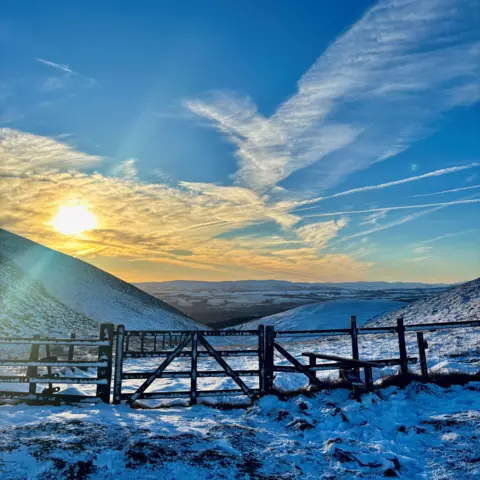 Lewis Almond Snowy ground in the foreground. A brown fence is in the middle of the image. A gate sits between two sections of the fence. Snowy hills are in the background. The sun is orange and glowing in the top right of the image. The sky is blue with white cloud streaked across it.