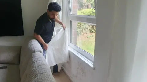 Man pulling back curtain with damp wall underneath a window